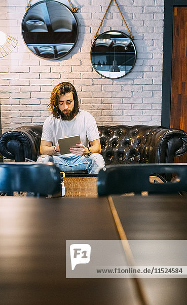 Stilvoller junger Mann sitzt auf der Couch in einem Cafe mit Tablette