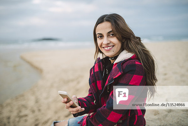 Porträt einer lächelnden jungen Frau mit Smartphone am Strand