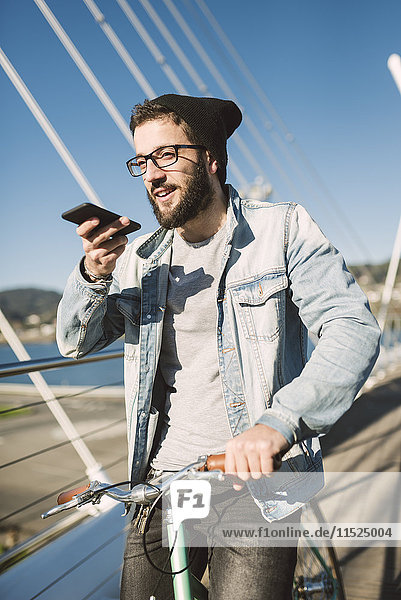 Junger Mann mit Fixie Bike auf einer Brücke mit Smartphone