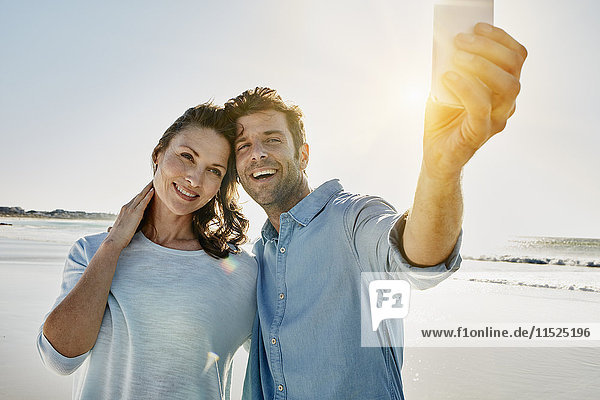 Porträt eines glücklichen Paares  das sich mit einem Smartphone am Strand aufhält.