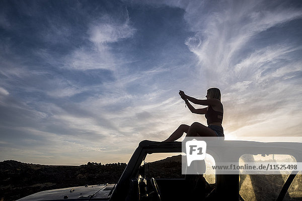 Silhouette of woman sitting on car roof at sunset taking picture