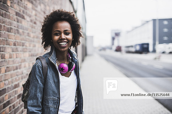 Porträt einer lächelnden jungen Frau mit Kopfhörer und Rucksack auf dem Bürgersteig stehend