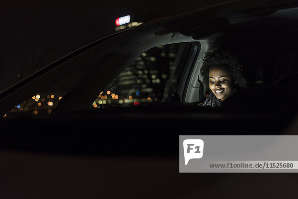Lächelnde junge Frau sitzt nachts in einem Auto mit Tablette