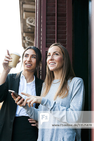 Two young women on balcony with cell phone and takeaway coffee looking up