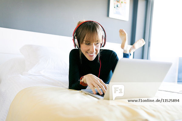 Lächelnde junge Frau auf dem Bett liegend  mit Laptop und Kopfhörer.