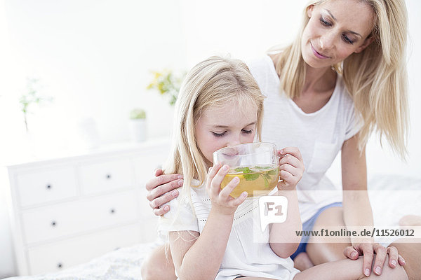 Mutter schaut der Tochter zu  wie sie Tee auf dem Bett trinkt.