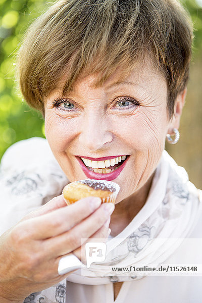 Porträt einer lächelnden älteren Frau  die einen Muffin im Freien isst.