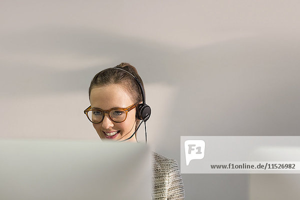 Smiling female telemarketer wearing headset talking on telephone at computer in office