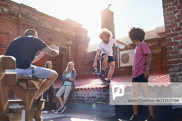 Mann mit Fotohandy fotografiert Freund beim Skateboard-Stunt auf dem sonnigen Stadtdach