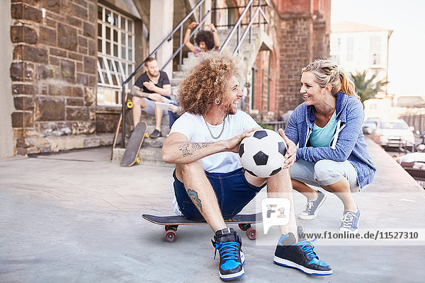 Lächelndes Paar mit Fußball und Skateboard im Gespräch auf dem städtischen Bürgersteig