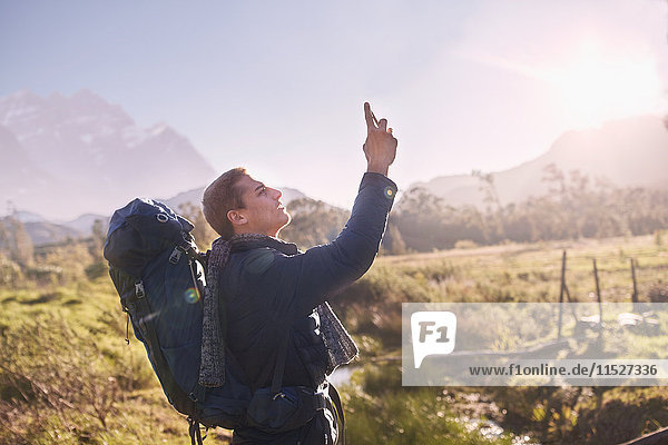 Young man with backpack hiking and using camera phone in sunny  remote field