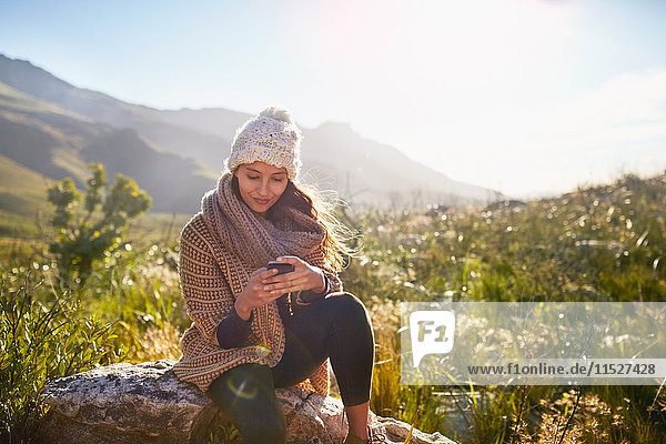Junge Frau SMS mit Handy auf Felsen im sonnigen  abgelegenen Feld