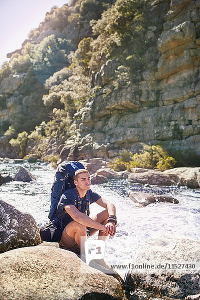 Junger Mann mit Rucksackwanderung  auf Felsen am sonnigen Fluss ruhend
