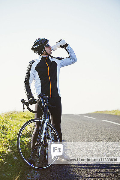 A cyclist by the side of a road  having a break and drinking from his water bottle.