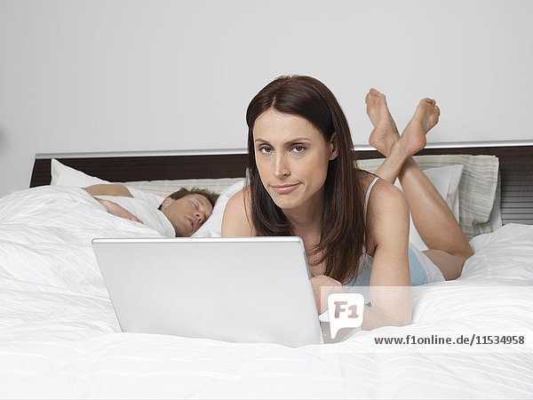Porträt einer Frau  die einen Laptop im Bett benutzt  während ein Mann schläft