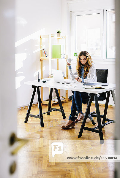 Junge Frau mit Tasse Kaffee und Smartphone am Schreibtisch im Home-Office