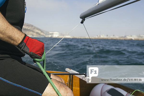 Hand mit rotem Handschuh eines Mannes  der ein Seil hält und mit seinem Segelboot auf See segelt.