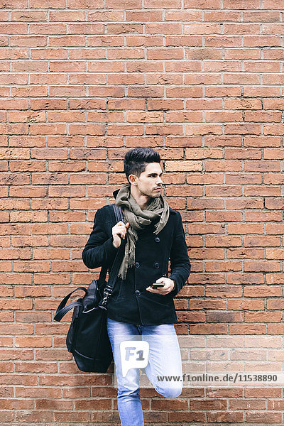 Young casual businessman leaning on brick wall carrying shoulder bag