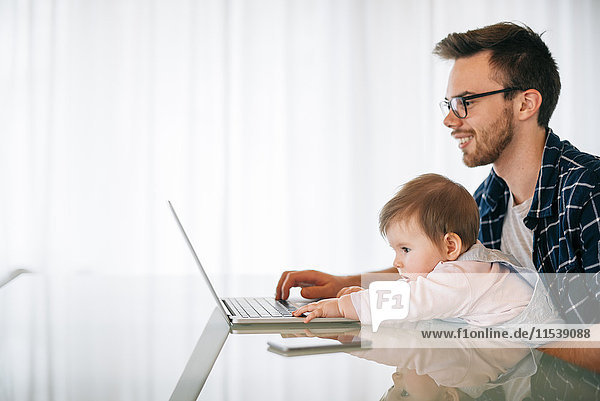 Junger Vater mit Baby auf dem Schoß mit Laptop