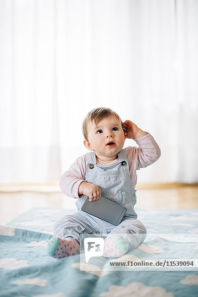 Portrait of baby girl sitting on blanket holding smartphone
