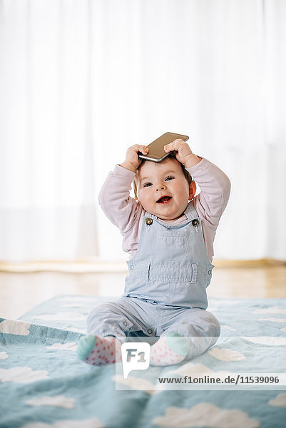 Portrait of baby girl sitting on blanket holding smartphone on the head