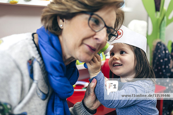 Porträt eines kleinen Mädchens  das das Ohr seiner Großmutter mit medizinischem Spielzeug untersucht.