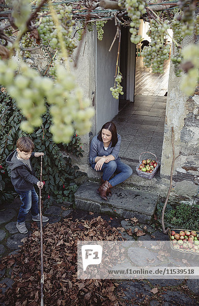 Spanien  Asturien  Frau sitzt am Eingang des Landhauses und beobachtet ihren Sohn beim Herbstlaubpflücken.