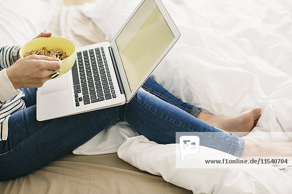 Frau auf dem Bett sitzend mit Laptop und Müslischale