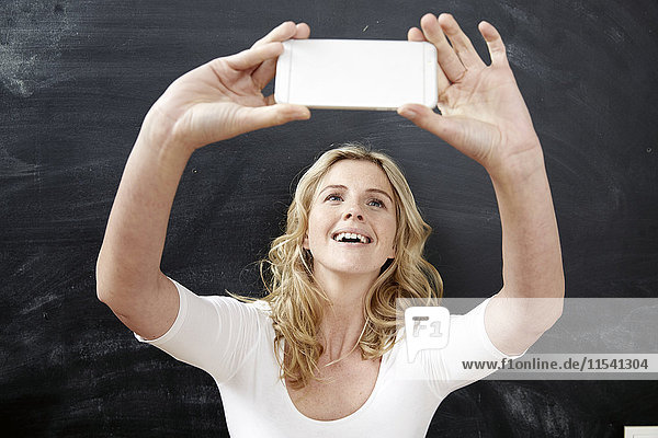 Porträt einer lächelnden jungen Frau  die einen Selfie mit Smartphone vor der Tafel nimmt.