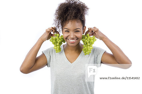 Portrait of smiling young woman holding green grapes like earrings in front of white background