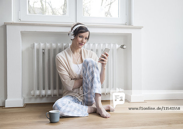 Frau zu Hause auf dem Boden sitzend mit Handy und Kopfhörer
