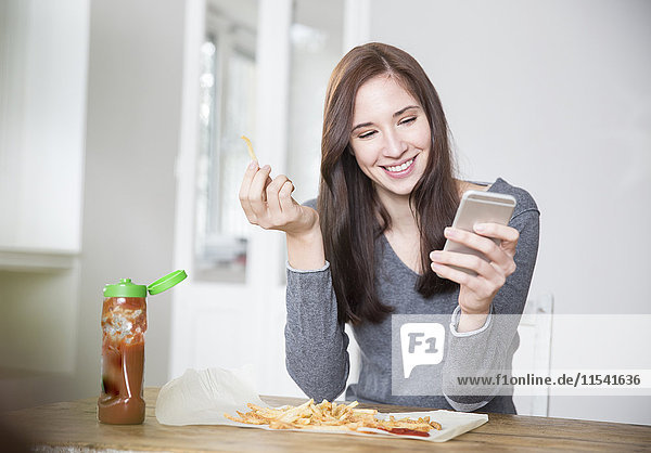 Porträt einer lächelnden jungen Frau mit Smartphone beim Essen von Pommes frites mit Ketchup