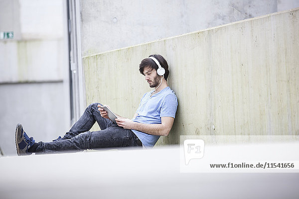 Junger Mann sitzt auf dem Boden und hört Musik mit Kopfhörern  die auf ein digitales Tablett schauen.
