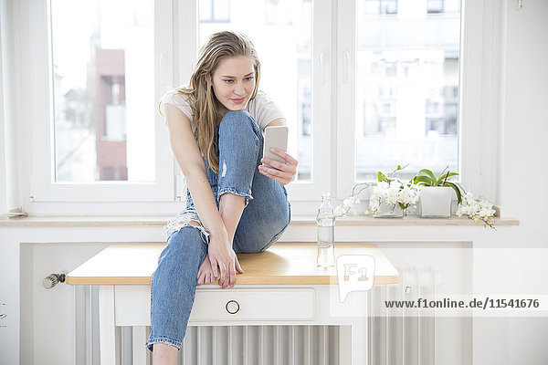 Junge Frau sitzt auf einem Tisch zu Hause und schaut auf das Smartphone.