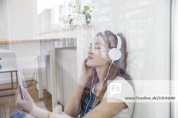 Junge Frau sitzt hinter der Fensterscheibe und hört Musik mit Kopfhörern.