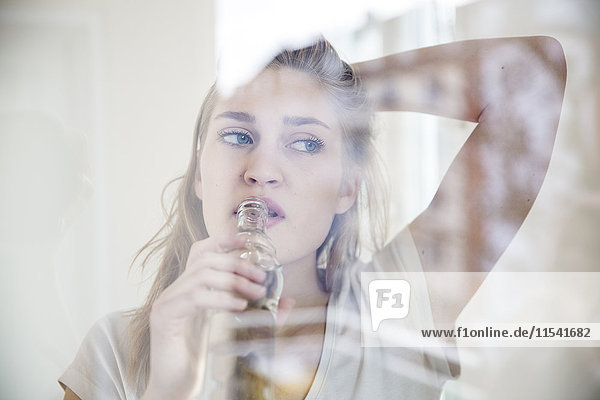 Porträt einer jungen Frau  die beim Trinken durch die Fensterscheibe schaut