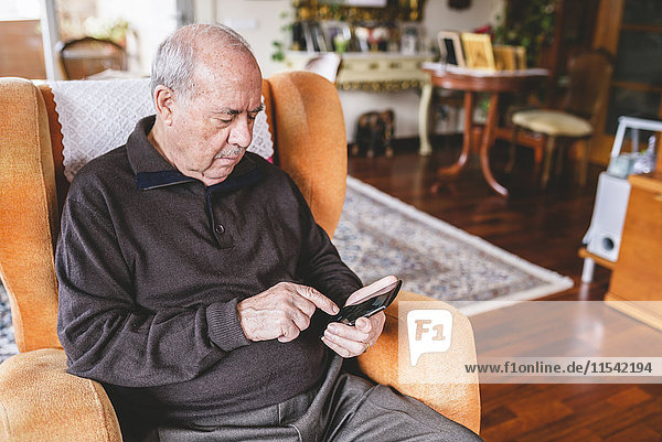 Senior Mann sitzt auf dem Sessel zu Hause mit dem Smartphone