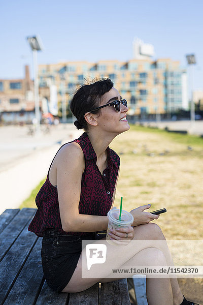 USA  New York City  Brooklyn  junge Frau sitzt auf einer Bank und hält Smartphone und Plastikbecher.