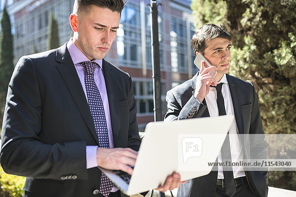 Two businessmen outdoors with laptop and cell phone