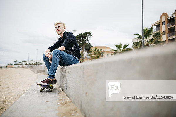 Spain  Torredembarra  smiling young skateboarder sitting on a wall