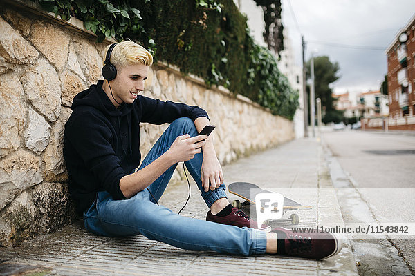 Spanien  Torredembarra  junger Skateboarder auf dem Bürgersteig sitzend Musik hören mit Kopfhörern