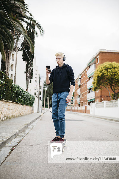 Spanien  Torredembarra  junger Mann mit Kopfhörern auf dem Skateboard und Blick auf sein Smartphone