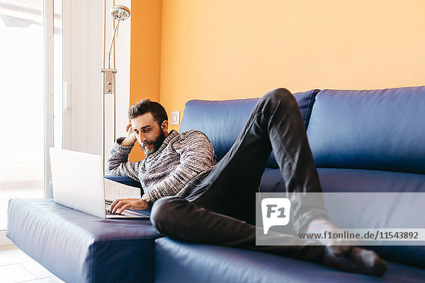 Bärtiger junger Mann arbeitet zu Hause entspannt auf der Couch liegend  mit Laptop