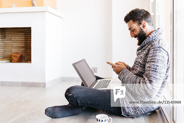 Der bärtige junge Mann arbeitet entspannt zu Hause  sitzt auf dem Boden  benutzt Laptop und Handy.