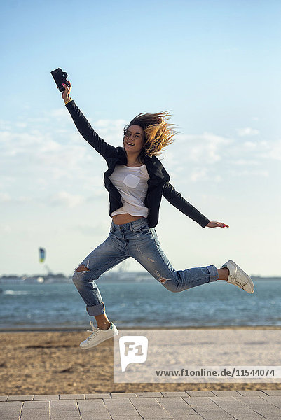 Spanien  Puerto Real  Frau mit Kamera springt in die Luft vor dem Meer