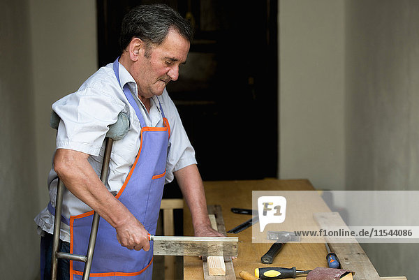 Senior man with crutch sawing piece of wood on workbench