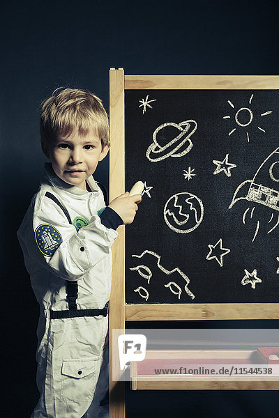 Kind mit Astronautenanzug zeichnet Himmelskörper und Leuchten auf eine Tafel