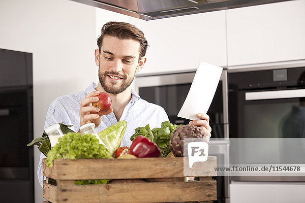 Porträt eines jungen Mannes mit Holzkiste mit frischem Gemüse und Einkaufsliste in seiner Küche