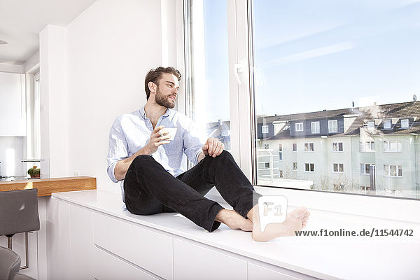 Young man with coffee cup sitting on sideboard at home looking through window