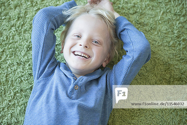 Porträt eines fröhlichen kleinen blonden Jungen auf grünem Teppich liegend